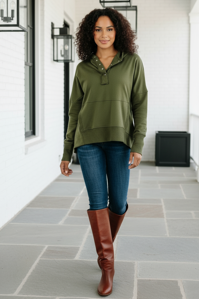 Woman wearing an olive green pullover, blue jeans, and brown knee-high boots in a hallway.